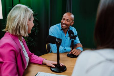 Profesyonel bir podcast stüdyosunda, mikrofonlar ve kameralarla yayın yapan, dinamik bir konuşma yapan üç kişiden oluşan çeşitli bir grup.