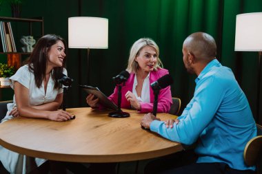 Bir grup insan bir masanın etrafında oturuyor, aktif olarak bir podcast kaydına bağlanıyor, bir kahkaha paylaşıyor ve profesyonel bir stüdyo ortamında içerik yaratıyorlar.