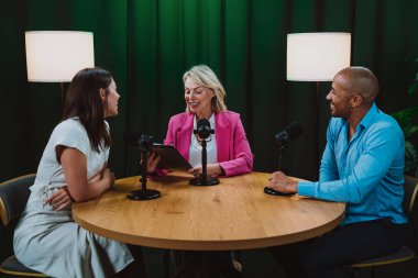 Bir grup insan bir masanın etrafında oturuyor, aktif olarak bir podcast kaydına bağlanıyor, bir kahkaha paylaşıyor ve profesyonel bir stüdyo ortamında içerik yaratıyorlar.