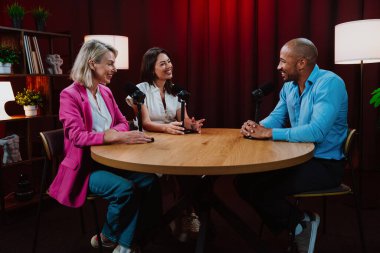 Profesyonel bir podcast stüdyosunda, mikrofonlar ve kameralarla yayın yapan, dinamik bir konuşma yapan üç kişiden oluşan bir grup.