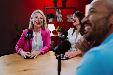 Profesyonel bir podcast stüdyosunda, mikrofonlar ve kameralarla yayın yapan, dinamik bir konuşma yapan üç kişiden oluşan bir grup.