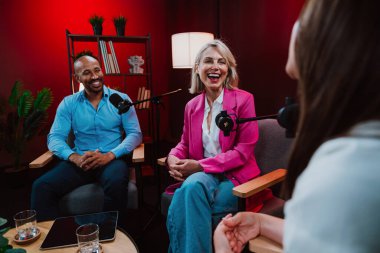 Profesyonel bir podcast stüdyosunda, mikrofonlar ve kameralarla yayın yapan, dinamik bir konuşma yapan üç kişiden oluşan bir grup.