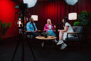 Profesyonel bir podcast stüdyosunda, mikrofonlar ve kameralarla yayın yapan, dinamik bir konuşma yapan üç kişiden oluşan bir grup.