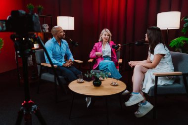 Profesyonel bir podcast stüdyosunda, mikrofonlar ve kameralarla yayın yapan, dinamik bir konuşma yapan üç kişiden oluşan bir grup.