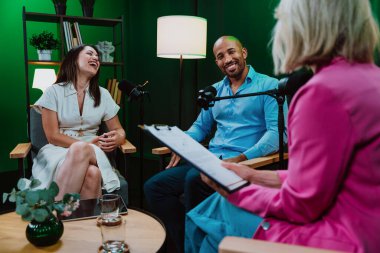 Profesyonel bir podcast stüdyosunda, mikrofonlar ve kameralarla yayın yapan, dinamik bir konuşma yapan üç kişiden oluşan bir grup.