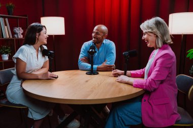 Bir grup insan bir masanın etrafında oturuyor, aktif olarak bir podcast kaydına bağlanıyor, bir kahkahayı paylaşıyor ve profesyonel bir stüdyo ortamında içerik yaratıyorlar.