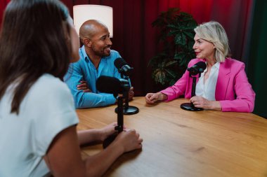 Bir grup insan bir masanın etrafında oturuyor, aktif olarak bir podcast kaydına bağlanıyor, bir kahkahayı paylaşıyor ve profesyonel bir stüdyo ortamında içerik yaratıyorlar.