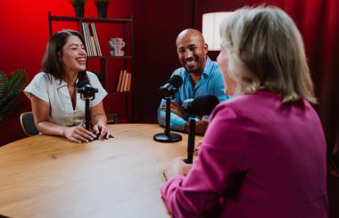 Bir grup insan bir masanın etrafında oturuyor, aktif olarak bir podcast kaydına bağlanıyor, bir kahkahayı paylaşıyor ve profesyonel bir stüdyo ortamında içerik yaratıyorlar.