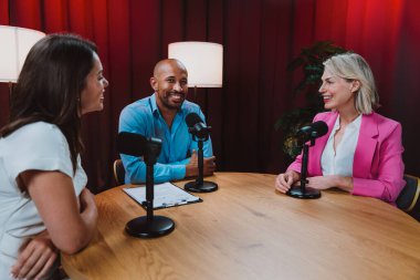 Üç farklı yetişkin sunucu, bir adam ve iki kadın, mikrofonlu bir masanın etrafında oturmuş, canlı bir podcast tartışmasına girişiyor ya da perdeli bir stüdyo ortamında röportaj yapıyorlar.