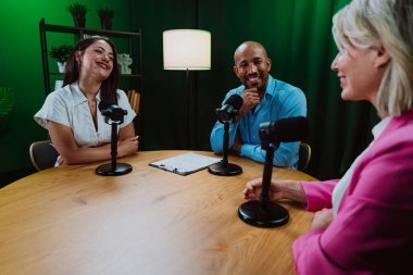 Üç farklı yetişkin sunucu, bir adam ve iki kadın, mikrofonlu bir masanın etrafında oturmuş, canlı bir podcast tartışmasına girişiyor ya da perdeli bir stüdyo ortamında röportaj yapıyorlar.