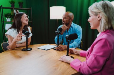 Üç farklı yetişkin sunucu, bir adam ve iki kadın, mikrofonlu bir masanın etrafında oturmuş, canlı bir podcast tartışmasına girişiyor ya da perdeli bir stüdyo ortamında röportaj yapıyorlar.