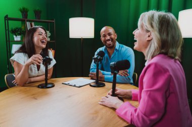 Üç farklı yetişkin sunucu, bir adam ve iki kadın, mikrofonlu bir masanın etrafında oturmuş, canlı bir podcast tartışmasına girişiyor ya da perdeli bir stüdyo ortamında röportaj yapıyorlar.