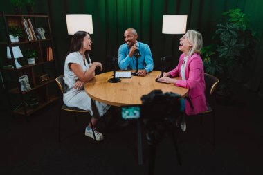 Üç farklı yetişkin sunucu, bir adam ve iki kadın, mikrofonlu bir masanın etrafında oturmuş, canlı bir podcast tartışmasına girişiyor ya da perdeli bir stüdyo ortamında röportaj yapıyorlar.