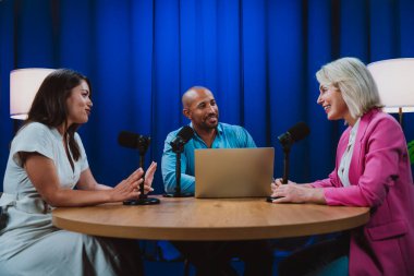 Üç sunucudan oluşan bir grup canlı bir ses yayını yakalıyor, mikrofonlar ve profesyonel bir stüdyoda dizüstü bilgisayarla eğlenceli bir konuşma yapıyorlar.