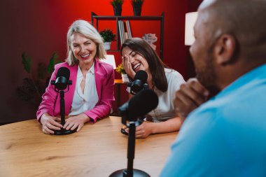 Profesyonel bir podcast stüdyosunda, mikrofonlar ve kameralarla yayın yapan, dinamik bir konuşma yapan üç kişiden oluşan bir grup.