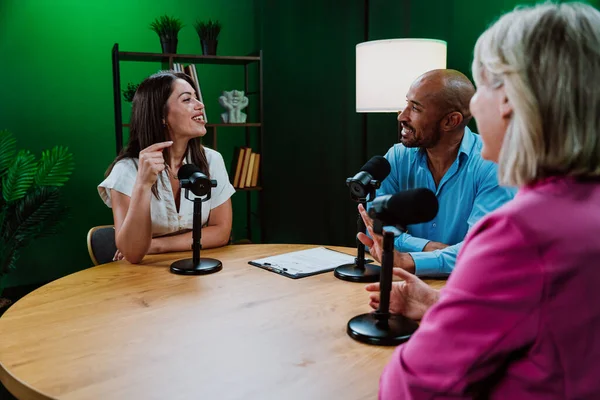 Üç farklı yetişkin sunucu, bir adam ve iki kadın, mikrofonlu bir masanın etrafında oturmuş, canlı bir podcast tartışmasına girişiyor ya da perdeli bir stüdyo ortamında röportaj yapıyorlar.