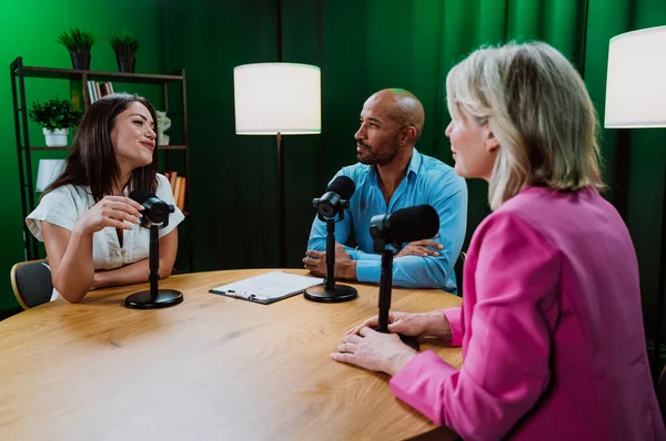 Üç farklı yetişkin sunucu, bir adam ve iki kadın, mikrofonlu bir masanın etrafında oturmuş, canlı bir podcast tartışmasına girişiyor ya da perdeli bir stüdyo ortamında röportaj yapıyorlar.