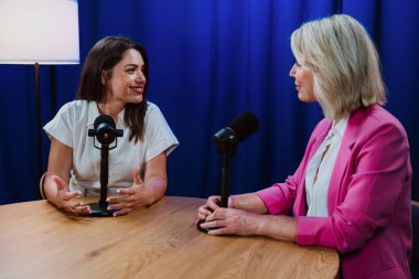 İki gülümseyen kadın bir podcast kaydediyor, mikrofonlara konuşuyorlar ve mikrofonlara konuşuyorlar profesyonel bir stüdyonun ahşap masasında mavi arka planı ve lambaları var.