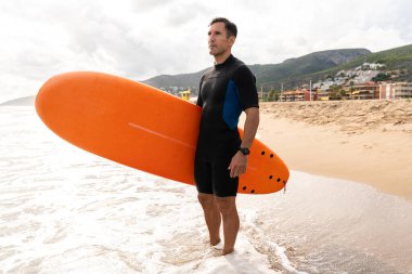 Dalgıç kıyafeti giymiş bir adam kumlu sahilde duruyor, sörf tahtası tutuyor, dalgaların kıyı şeridinde, dağların ardında dalgalanacağı bir sörf sezonu bekliyor.