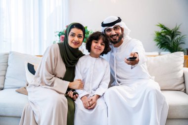 Geleneksel emirati kıyafetleri giyen Ortadoğu ailesi evde birlikte vakit geçiriyorlar. Arap aileler gülümsüyor, televizyon seyrediyor ve kanepede dinleniyorlar.