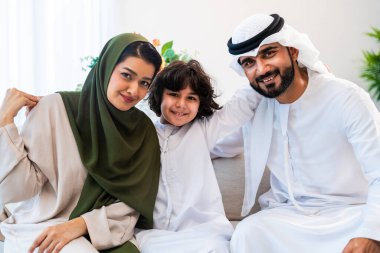 Geleneksel emirati kıyafetleri giyen Ortadoğu ailesi evde birlikte vakit geçiriyorlar. Orta Doğulu ebeveynler ve çocuklar birlikte gülümsüyor, beraberliği ve aile sevgisini temsil ediyor.