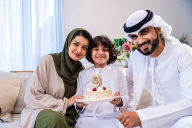 Geleneksel emirati kıyafetleri giyen Ortadoğu ailesi evde birlikte vakit geçiriyorlar. Orta Doğulu aile gülümsüyor ve elinde bir doğum günü pastası tutuyordu.