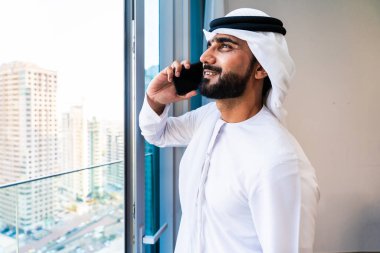 Yüksek katlı bir apartmanın penceresinden dışarı bakan, cep telefonuyla mutlu bir şekilde konuşan, Dubai 'de başarılı ve bağlı bir şehir yaşam tarzını benimseyen, kendinden emin emirati bir adam.