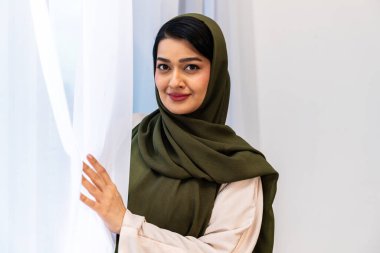 Yeşil tesettürlü ve açık renk elbiseli Emirati kadını, güneşli bir pencerenin yanında güvenle gülümsüyor. Saf perdelerle, huzur, modern ev sıcaklığı ve huzur getiriyor.