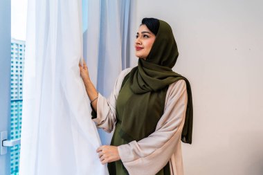 Abaha 'daki Emirati kadını ve tesettür pencerenin yanında duruyor, beyaz perdeleri açıyor, sakin ve umut dolu bir ifade dışarıdan modern şehir binalarına bakarken