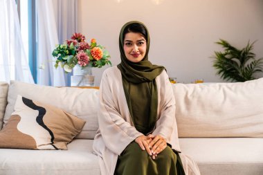 Genç Arap kadın tesettürlü ve abaya, evinde modern bir koltukta rahatça oturuyor, günlük hayatta mütevazı ve kültürel kimliği temsil ediyor.
