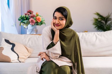 Emirati kadını tesettürlü ve abaya evdeki modern oturma odasındaki kanepede dinleniyor, sakin, şık bir ev portresiyle kameraya güvenle gülümsüyor.