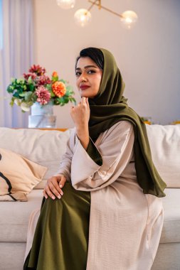Emirati kadını güzel dekore edilmiş çağdaş bir oturma odasında rahat bir kanepede oturuyor, geleneksel bir abaya ve tesettür giyiyor, kameraya doğru güvenli bir şekilde bakıyor