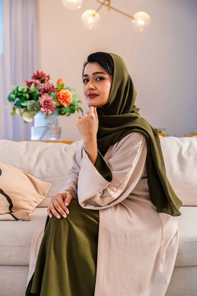 Emirati kadını güzel dekore edilmiş çağdaş bir oturma odasında rahat bir kanepede oturuyor, geleneksel bir abaya ve tesettür giyiyor, kameraya doğru güvenli bir şekilde bakıyor