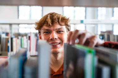 Genç bir erkek öğrenci kütüphane koridorunda kameraya gülümserken raftaki bir kitaba uzanıyor ve okumak ve öğrenmek için okuma materyali seçiyor.