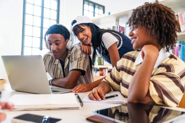 Kütüphanede toplanan çok ırklı bir grup genç öğrenci - modern üniversite kütüphanesinde dizüstü bilgisayar ve ders kitapları kullanarak araştırma projesinde işbirliği yapan çeşitli üniversite öğrencileri