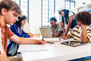 Kütüphanede toplanan çok ırklı bir grup genç öğrenci. Çok ırklı genç öğrenciler kütüphane masasında, dizüstü bilgisayarlar, tabletler ve not defterleri kullanarak iş birliği yapıyorlar.