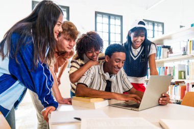 Kütüphanede bir araya gelen çok ırklı genç öğrenci grubu. Modern bir kütüphanede bir dizüstü bilgisayara bakıp gülümseyen ve işbirliği yapan çok ırklı genç öğrenciler.