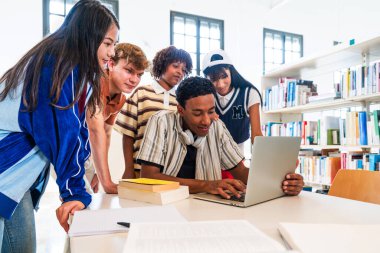 Kütüphanede toplanan çok ırklı bir grup genç öğrenci - bir kütüphanede proje üzerinde çalışan genç öğrenciler, çalışmak ve araştırmak için dizüstü bilgisayar ve ders kitapları kullanıyorlar.