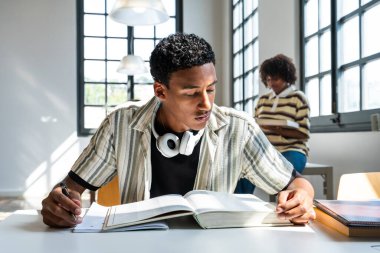 Ders kitapları ve okul malzemeleriyle çevrili bir kitap okumaya ve notlar yazmaya odaklanan genç adam, bir üniversite kütüphanesinde öğrenim ve eğitime olan bağlılığını yansıtıyor.