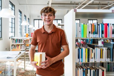 Genç bir erkek öğrenci gururla kitap tutarken gülümsüyor, parlak bir üniversite kütüphanesinde kitaplıkların arasında duruyor, eğitim ve bilgiyi temsil ediyor.