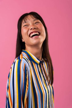 Renkli arka planda güzel bir Çinli kadın portresi. Neşeli genç Asyalı kadın geniş bir gülümseme, mutluluk ve neşe ifade ediyor, pembe arka planda renkli çizgili bir gömlek giyiyor.