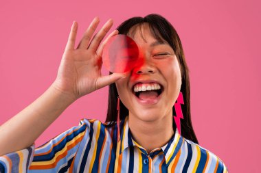 Renkli arka planda güzel bir Çinli kadın portresi. Canlı çizgili gömleği ve küpeleri olan neşeli Asyalı kadın kırmızı filtreden bakıp gülüyor. Pembe stüdyo arka planı