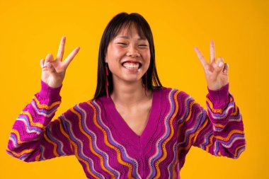 Renkli arka planda güzel bir Çinli kadın portresi. Asyalı kadın poz veriyor, geniş bir şekilde gülümsüyor ve canlı sarı stüdyo geçmişine karşı çift barış işareti gösteriyor.