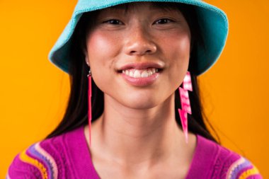 Renkli arka planda güzel bir Çinli kadın portresi. Stüdyoda turuncu arka planda poz veren genç Asyalı kadın, mavi kova şapka ve pembe yıldırım küpeleri takıyor.