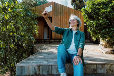 Yaşlı kadın kırsal ahşap merdivenlerde dinleniyor, düşünceli bir şekilde uzaklardan bakıyor, doğayla çevrili basit ahşap kulübesinde huzurlu bir emeklilik yaşam tarzını kucaklıyor.