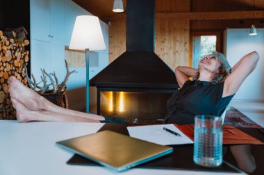 Olgun bir kadın ofis masasında oturuyor, bilgisayarla, belgelerle ve bir bardak suyla çalışıyor, profesyonel üretkenliği ve uzaktan çalışmayı temsil ediyor.