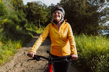 Kirli bir yolda dağ bisikletinin yanında duran, gülümseyen ve mesafeye bakan, manzaralı yeşil bir dağ zeminiyle aktif ve sağlıklı bir yaşam tarzını kucaklayan yaşlı bir kadın.