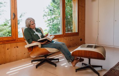 Üst düzey bir kadın evde rahatlıyor, rahat bir koltukta otururken kitap okuyor, büyük bir pencerenin yanında boş vaktinin keyfini çıkarıyor ve yanında bir dizüstü bilgisayarla bir sedirin üzerinde dinleniyor.