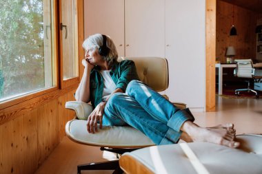 Deneyimli, gri saçlı bir kadın modern bir oturma odasında dinleniyor, kulaklık takıyor, koltukta çıplak ayakla oturuyor, müzik dinliyor ve dikkatlice pencereden dışarı bakıyor.