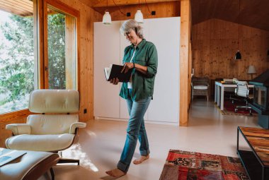 Kıdemli kadın çıplak ayakla duruyor, rahat bir yaşam alanında açık bir kitap okuyor, modern bir şömine ve ahşap duvarlı, boş ve bilgili bir anın tadını çıkarıyor.